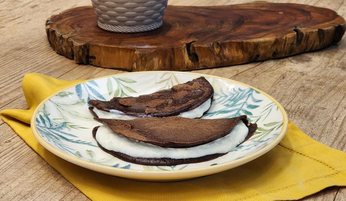 Fotografia em tons de marrom com um prato redondo ao centro. Em cima do prato existe duas panquecas de chocolate com aveia recheadas com um creme de iogurte.