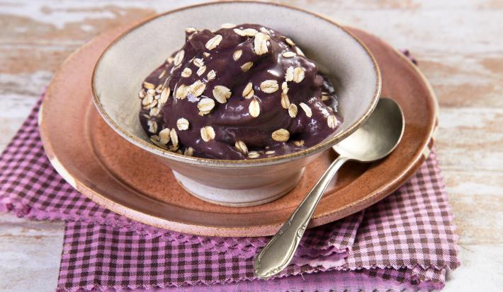 Fotografia em tons de roxo em uma bancada de madeira com um pano xadrez rosa e roxo, um prato raso rosa, um bowl fundo com o açaí dentro dele.