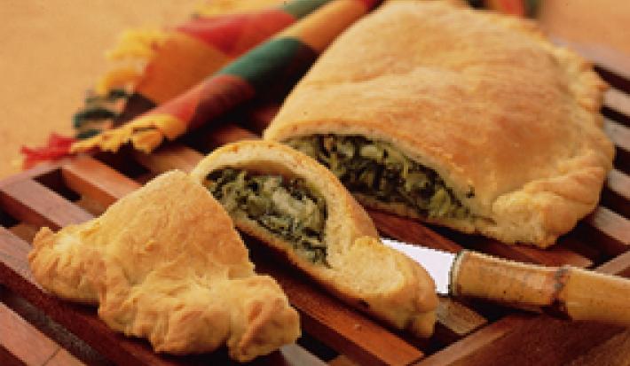 Fotografia em tons de amarelo em uma mesa de madeira com um pano colorido, uma tábua de madeira colorida com o calzone recheado de escarola cortado ao meio