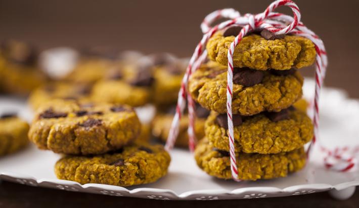 Fotografia em tons de amarelo em uma bancada de madeira com um recipiente branco raso e vários cookies integrais de aveia e abóbora em cima dele.