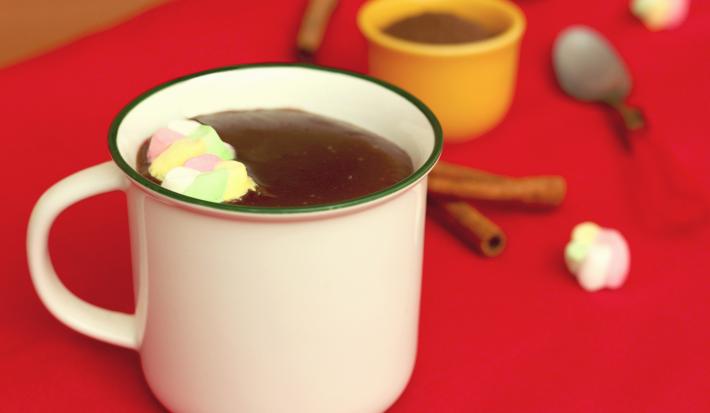 Fotografia em tons de branco e vermelho, ao centro uma xícara branca com chocolate quente e marshmallows, sobre toalha vermelha. Atrás um potinho com achocolatado e uma colher, com paus de canela e marshmallows espalhados ao redor.