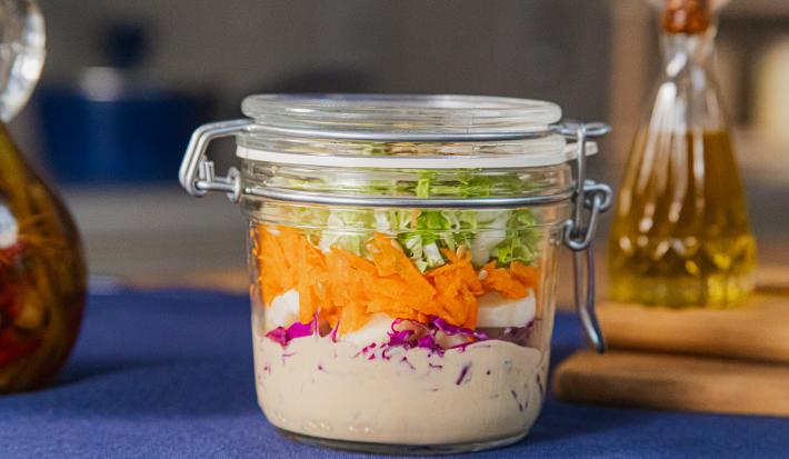 Fotografia em tons de azul com um pote de vidro ao centro. Dentro do pote existe uma receita de salada de repolho roxo feita com repolho, cenoura, palmito e molho rosé