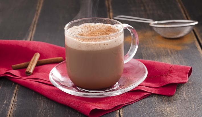 Fotografia em tons de cinza e rosa de uma bancada de madeira escura, um pano rosa escuro com duas canelas em pau, um pires de vidro e sobre ele uma xícara transparente com o cappuccino. Ao fundo uma peneira com canela em pó.