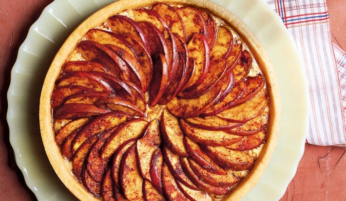 Foto tirada de cima de uma torta redonda de maça em cima de um prato branco. A torta é decorada por várias maças cortadas.