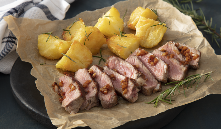 Foto em tons escuros da receita de bife ancho com batatas servidaem tiras de bife sobre um papel marrom em cima de uma pedra toda preta. Ao fundo há um pano branco quadriculado e ramos de alecrim