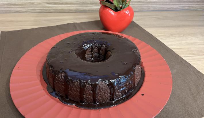 Fotografia de um bolo de chocolate e café com calda de chocolate sobre um apoio grande vermelho. O apoio está por cima de um pano marrom de tom escuro.