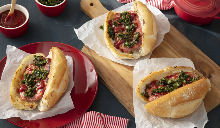Foto em tons de vermelho da receita de choripan tradicional servida em três porções, duas sobre uma tábua de madeira e uma sobre um prato vermelho. Ao fundo há dois bowls de porcelana vermelhos além de uma panelinha e um paninho vermelhos