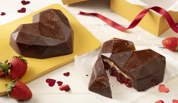 Fotografia em tons de marrom, amarelo e vermelho de uma bancada com dois corações de chocolate, um deles partido ao meio mostrando o recheio branco e de geleia de morango. Ao redor morangos e corações decorando.