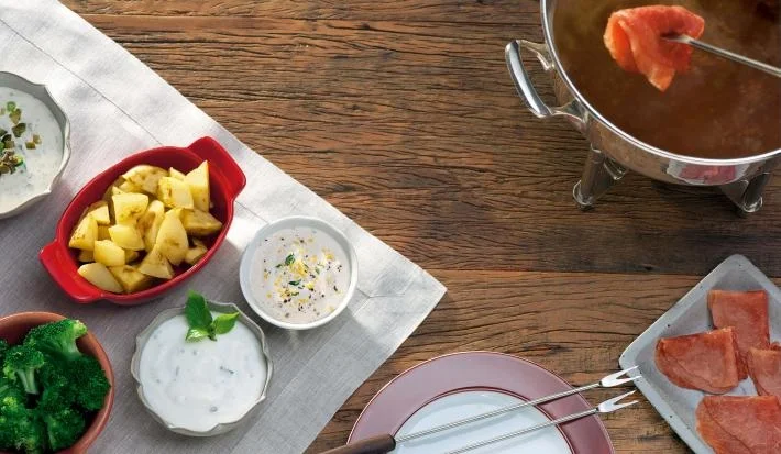 Imagem de uma mesa de madeira com fondue de carne com acompanhamentos e molhos.