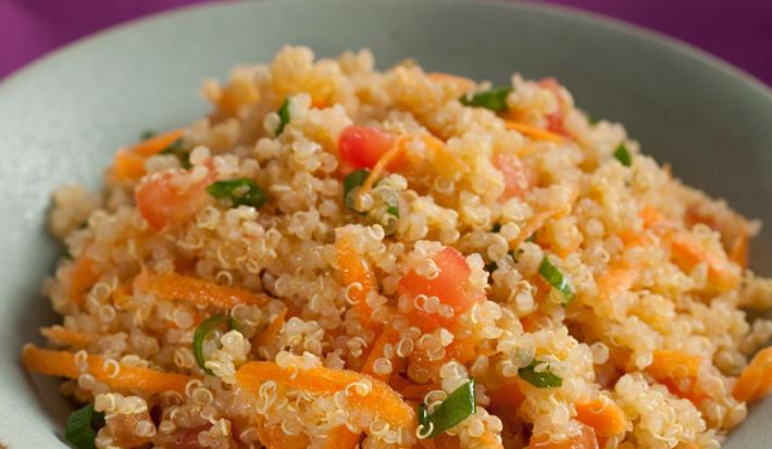 Fotografia em tons de laranja de uma bancada roxa com um prato fundo cinza com a salada de quinoa, tomate, cenoura ralada e cebolinha.
