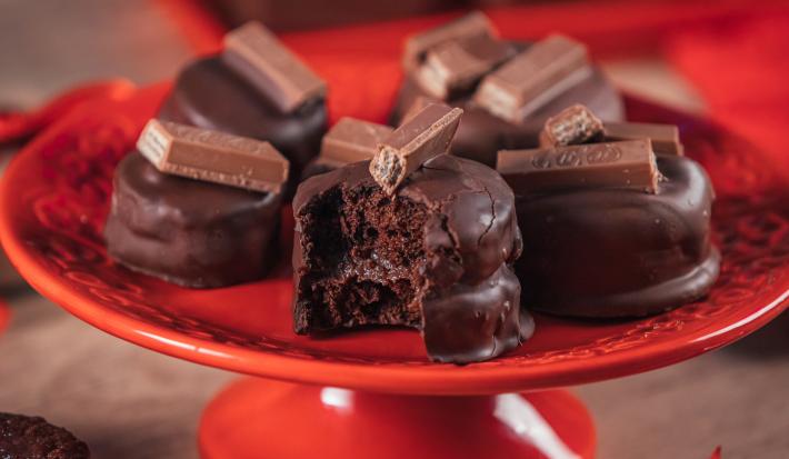Fotografia em tons de vermelho em uma bancada de madeira e um prato vermelho redondo ao centro. Em cima dele, existem pães de mel de chocolate com KitKat em cima decorando e um aberto mostrando o recheio.