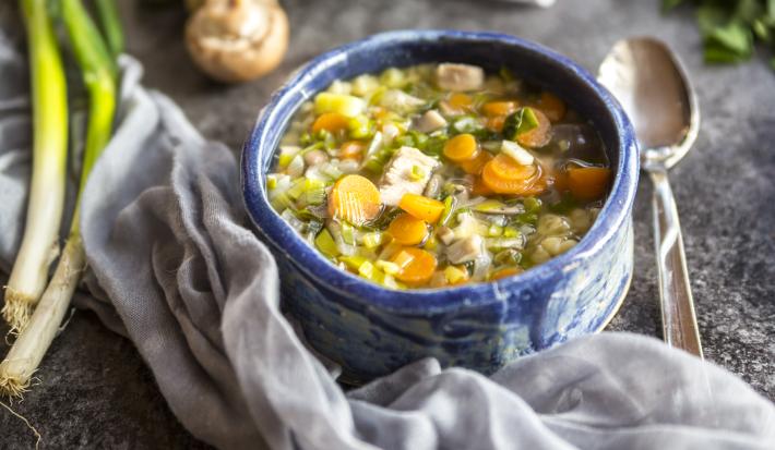 Fotografia em tons claros, no centro, um recipiente de vidro na cor azul. Dentro, uma sopa com cenoura, alho-poró e mandioquinha. Ao lado, uma colher de sopa, um alho-poró sobre a mesa. No fundo da foto, alguns champignons sobre a mesa.