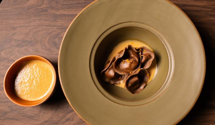 Fotografia vista de cima de um capeletti de cacau e café, recheado com creme de banana, por cima de um creme anglaise de café. A receita está em um prato fundo de bordas grossas e cor verde, ao lado tem um recipiente fundo pequeno com mais molho.
