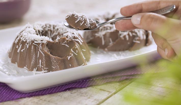 .Fotografia em tons de roxo e branco de uma bancada de madeira branca com um paninho roxo, sobre ele um prato retangular com dois gelados de dark chocolate. Sobre ele uma colher com um pedaço do gelado. Ao fundo um recipiente redondo com coco.