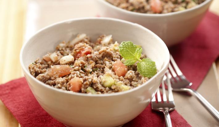 Fotografia em tons de vermelho em uma mesa de madeira clara com uma tábua de madeira, dois guardanapos de papel vermelhos e dois recipientes redondos com a salada de trigo com carne moída.