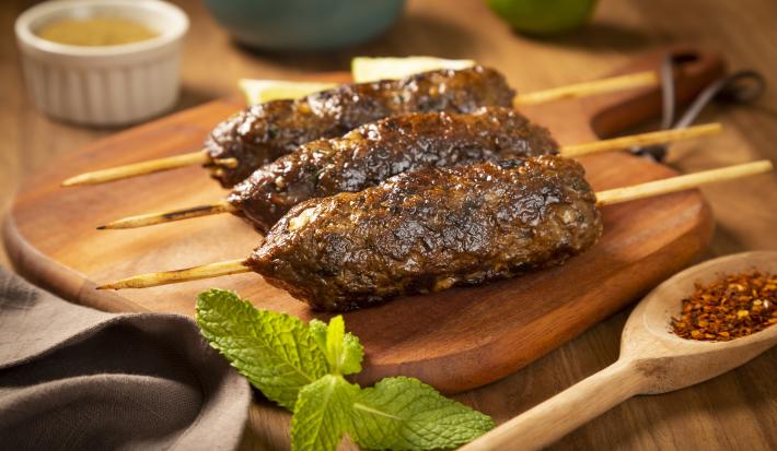 Fotografia em tons de marrom, verde e cinza de uma bancada de madeira marrom vista de frente. Uma tábua de madeira contém 3 espetinhos com carne moída, ao lado uma colher de madeira com tempero, um pano cinza e um potinho redondo branco com tempero.