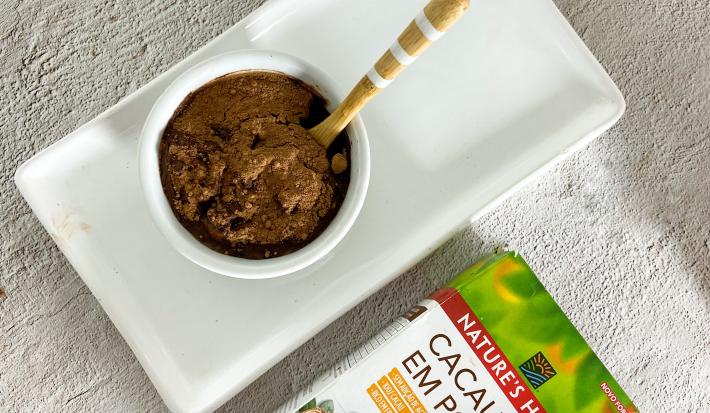 Foto em tons claros de cinza da receita de mousse de cacau com 3 ingredientes servida em um bowl de porcelana branco sobre uma base de porcelana de mesma cor. Ao lado, há uma embalagem de cacau em pó nature's heart