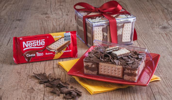 Foto da receita de Cake Box de Paçoca com Choco Trio. Observa-se duas caixas de acrílico com a receita dentro sobre um prato vermelho quadrado e um pano de prato amarelo.
