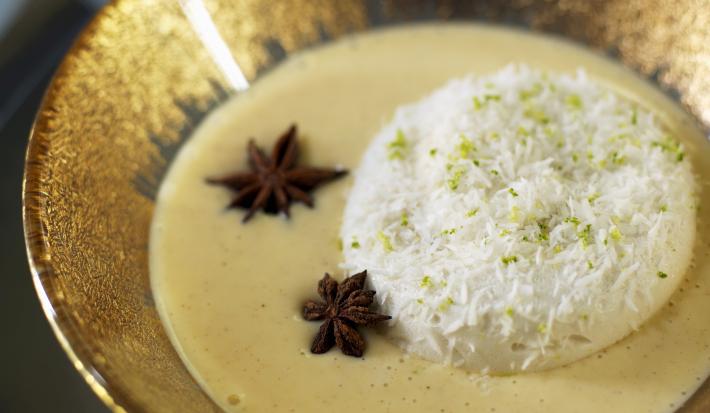 Fotografia de ovos nevados com um creme inglês, algumas raspas de limão por cima e dois anis estrelados no creme. A sobremesa está dentro de um recipiente fundo de alumínio na cor dourado, que está sobre uma mesa preta.