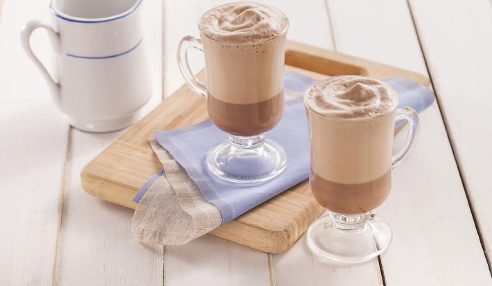 Fotografia em tons de azul em uma bancada de madeira clara, uma tábua de madeira, um paninho azul e duas xícaras de vidro com o cappuccino gelado de cacau dentro deles. Ao fundo, uma jarra branca com azul com mais cappuccino.