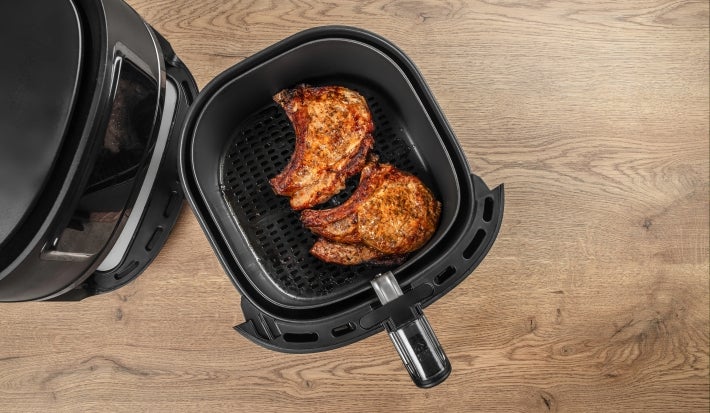Fotografia vista de cima uma air fryer preta, com a cesta afastada sobre uma mesa de madeira clara. Dentro da Air Fryer tem duas bistecas de porco douradinhas.