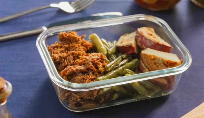 A fotografia mostra uma marmita de vidro com carne desfiada dourada, batata-doce assada levemente crocante e vagem refogada vibrante, organizada de forma prática e equilibrada, com cores intensas e aparência convidativa.