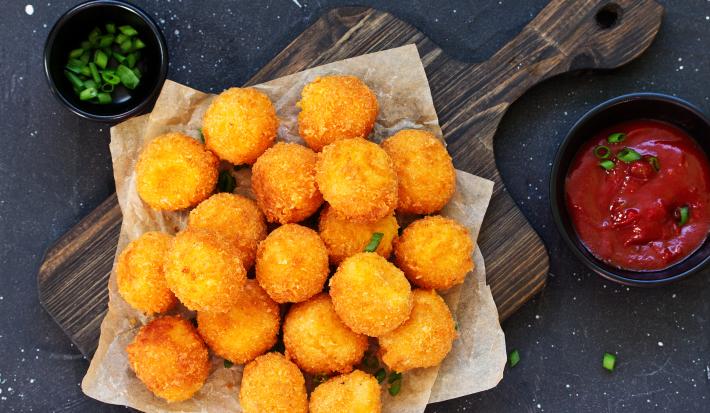 Fotografia em tons de amarelo e marrom com uma tábua ao centro. Em cima da tábua existe pequenas bolinhas fritas acompanhadas com um pequeno pote de ketchup.