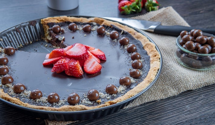 Fotografia de uma torta grande de bombom de morango dentro de uma forma redonda preta. Por cima da ganache da torta temumas bolinhas de chocolate e morangos em pedaços decorando-a. Ao lado tem uma faca e mais bolinhas de chocolate num potinho.