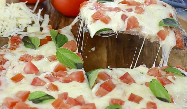 Foto aproximada de uma pizza marguerita, decorada com tomates e folhas de manjericão, sobre uma tábua de madeira e com tomates ao fundo