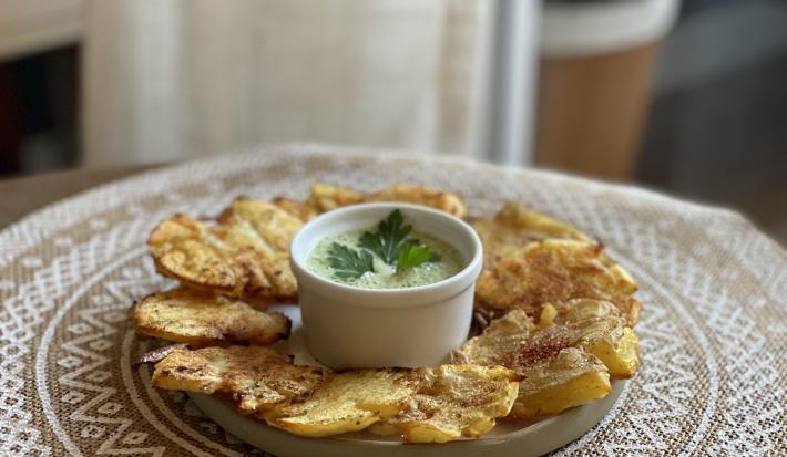Fotografia em tons de marrom em uma bancada de madeira com uma toalha redonda branca de renda e um prato bege em cima com as batatas assadas e temperadas com um potinho branco ao centro com o molho de maionese verde.