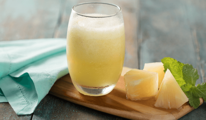 Fotografia em tons de verde em uma bancada de madeira mesclada, um pano verde água, uma tábua de madeira, um copo de vidro com a vitamina de abacaxi dentro, folhas de hortelã e pedaços de abacaxi ao lado.