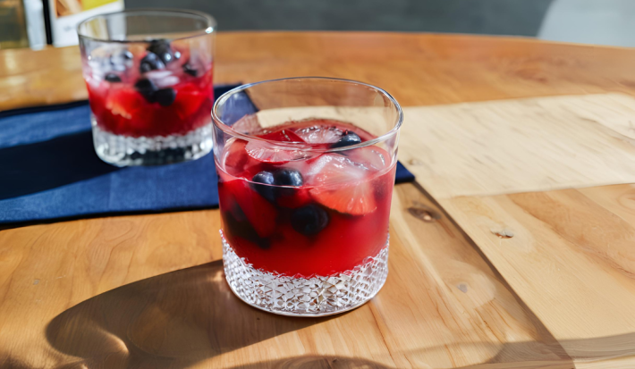 Fotografia de dois copos de vidro baixos com uma bebida sem álcool com frutas vermelhas e gelo. Os copos estão afastados um do outro, e o copo do fundo está sobre um pano azul de tom escuro, sobre uma mesa de madeira.