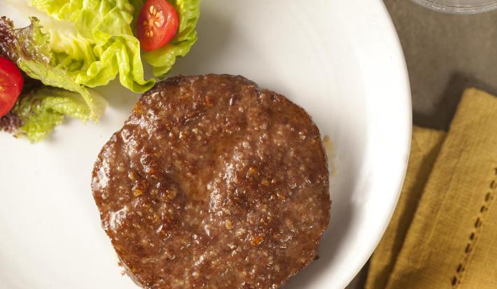 Fotografia em tons de amarelo em uma bancada de madeira, um pano amarelo, um prato redondo branco com salada de alface e tomate e ao lado uma carne de hambúrguer.