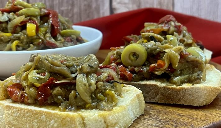 Foto da receita de caponata servida em duas fatias pão italiano e, atrás, em um bowl branco. Esses elementos estão sobre uma mesa de madeira redonda com um pano vermelho decorativo ao fundo