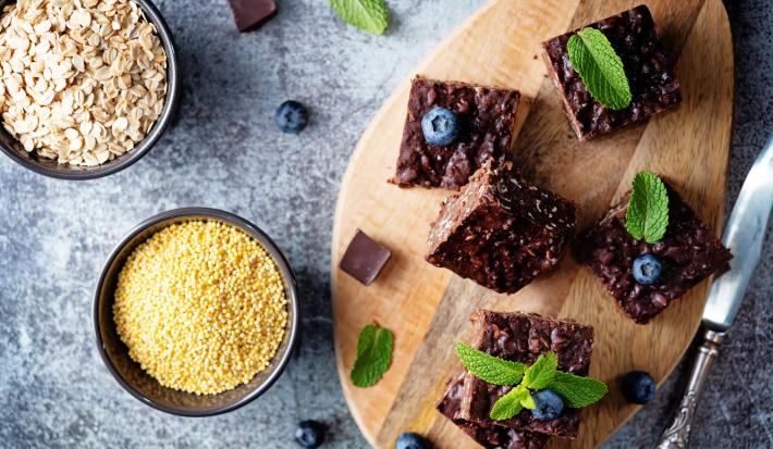 Fotografia em tons de marrom, cinza e amarelo em uma bancada de mármore cinza, uma tábua de madeira e em cima dela 5 brownies de chocolate. Ao lado, potinhos com farinha de aveia e uma faca.