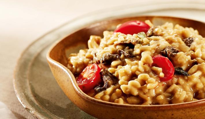 Fotografia em tons de vermelho em uma mesa com uma toalha bege, um prato verde raso grande, um bowl de madeira com o risoto de funghi com tomatinhos dentro dele.