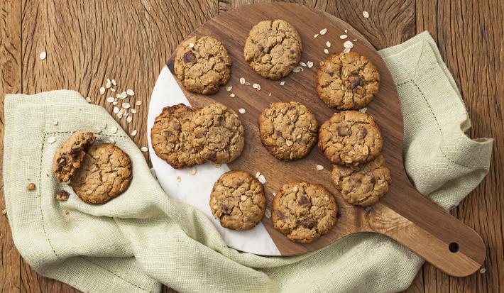 Fotografia em tons de marrom e bege, com tábua de cozinha ao centro com cookies assados e sobre guardanapo bege, tudo em bancada de madeira marrom com flocos de aveia espalhados.