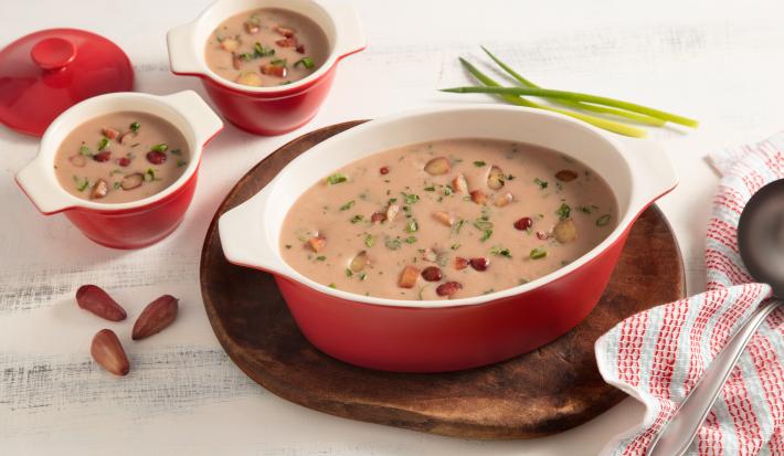 Fotografia em tons de branco e vermelho de uma bancada vista de cima. Em cima de uma tábua redonda contém um recipiente oval com alças nas cores vermelho e branco com a sopa e pedaços de pinhão por cima, ao lado dois potinhos com a sopa e um pano