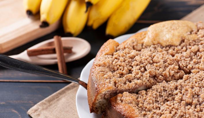 Fotografia uma cuca de banana com farofa sobre um prato de vidro branco raso, apoiado em um pano marrom de tom claro. Tem uma fatia da torta sendo retirada com uma espátula de prata. Ao lado tem uma colher branca com pau de canela e bananas inteiras.