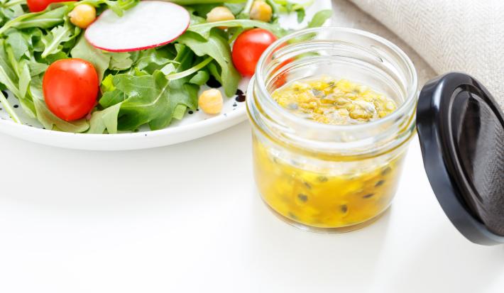 fotografia em tons de branco, verde e amarelo de uma bancada branca vista de cima. Contém um pote transparente com molho de maracujá e ao lado um prato redondo branco com salada.