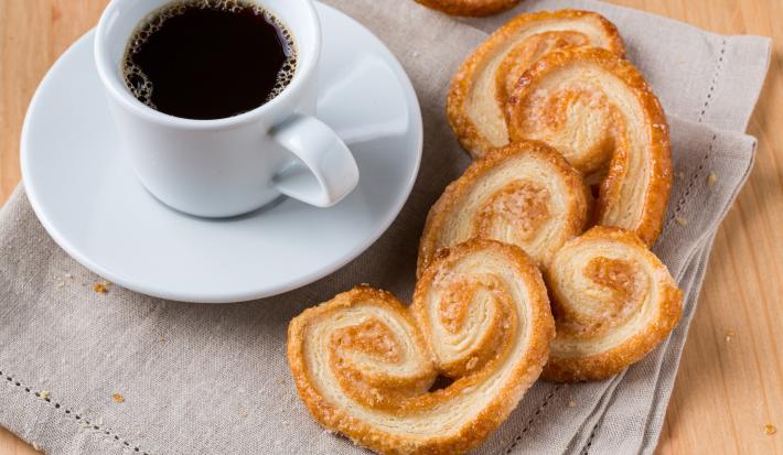 Foto da receita de Palmier ou Orelha de Macaco. Observa-se o biscoito em disposto em cima de um pano de prato e, ao lado, uma xícara com café sobre um pires.