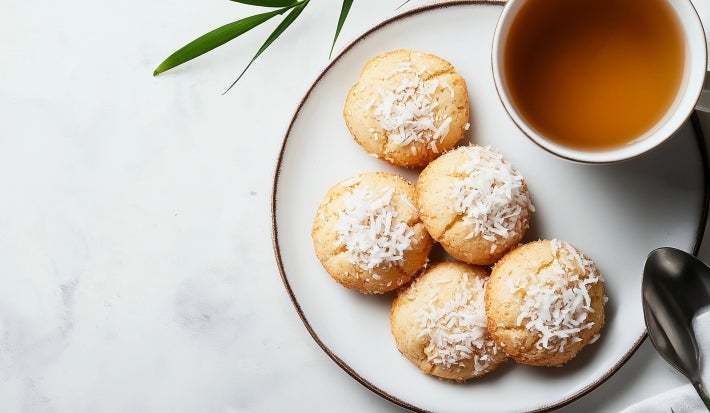 Em cima de uma mesa, prato redondo com biscoitos amanteigados, decorado com coco ralado. Ao lado direito, xícara de chá.