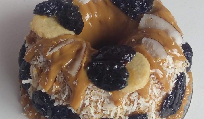Foto da receita de Bolo de tapioca com doce de leite e ameixa. Observa-se um bolo coberto de doce de leite com cocos grudados na lateral e ameixas decorando.