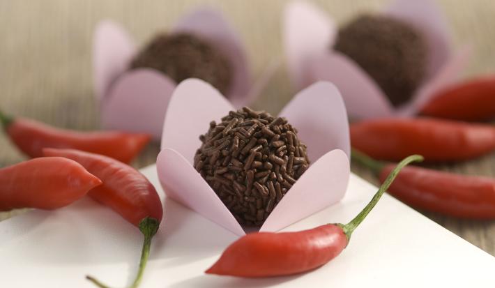 Fotografia em tons de vermelho em uma bancada de madeira com uma toalha branca, ao centro um brigadeiro em uma forminha rosa e várias pimentas vermelhas espalhadas pela mesa.