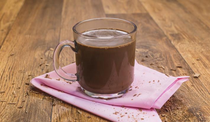 Fotografia de uma bancada de madeira com um paninho rosa e uma xícara transparente com o chocolate quente dentro.