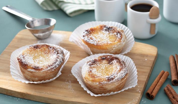 Fotografia em tons de amarelo em uma bancada de madeira de cor azul. Ao centro, uma tábua de madeira contendo os pasteis. Ao lado, há uma peneira de inox e ao fundo, duas xícaras de café.