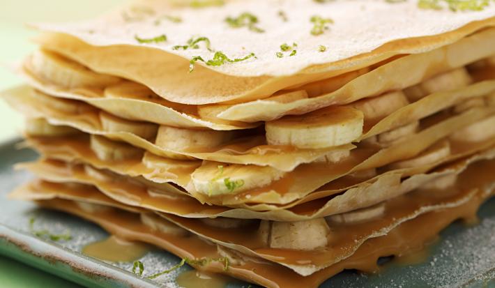 Fotografia em tons de verde em uma bancada de madeira com uma toalha verde, um prato quadrado com a torta crocante com recheio de doce de leite e bananas em rodelas.