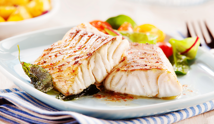 Fotografia mostra dois filés de peixe grelhado sobre um prato branco e salada.