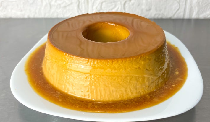 Fotografia em tons claros com um pudim de doce de leite ao centro. O pudim possui uma cor forte de doce de leite e está coberto com uma calda dourada