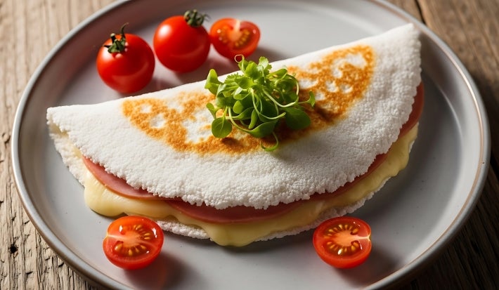 Fotografia em tons claros de uma tapioca recheada com queijo, presunto, orégano e alguns tomatinhos em volta, dentro de um prato cinza de vidro raso.